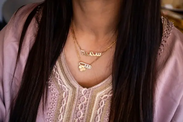 A close-up of a woman wearing two layered, personalized gold-plated nameplate necklaces over an embroidered pink top.