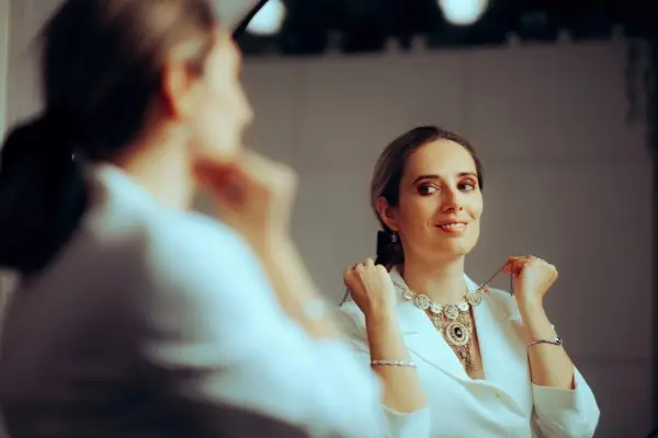 A woman smiles at her reflection in a mirror while trying on an ornate, gold-plated statement necklace over a white blazer.