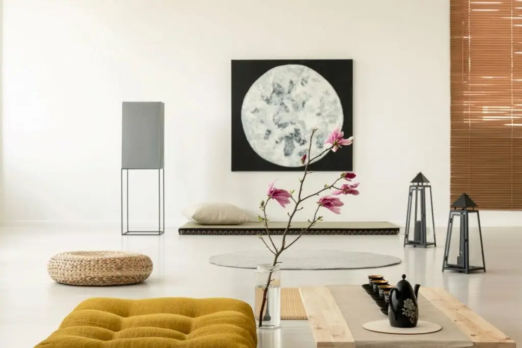 A serene and minimalist modern Japanese living room featuring a low-profile grey sofa, a simple round wooden coffee table, and a large window with a view of green foliage, embodying Wabi-sabi principles.