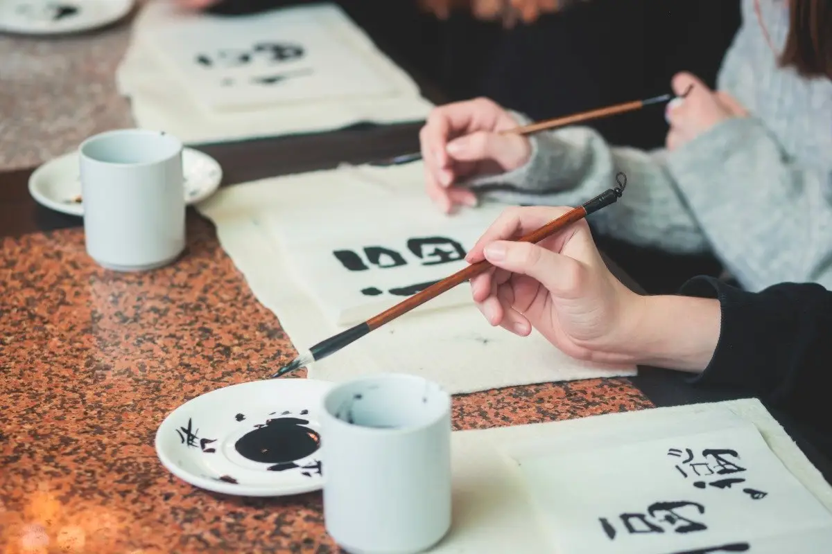 A top-down view of people practicing Japanese calligraphy (Shodo), with various tools like brushes, an ink stone, and rice paper laid out on a table as characters are being painted.