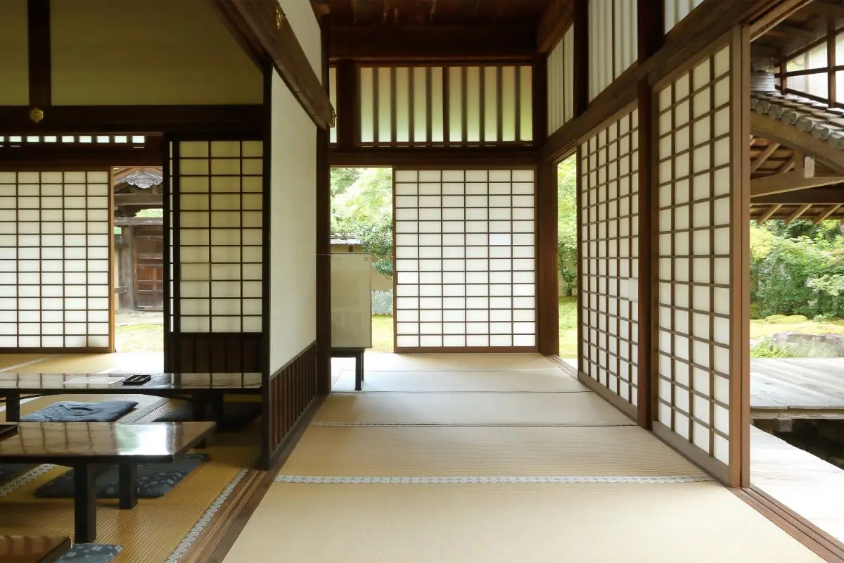 A tradtional Japanese home interior showcasing natural materials, with light wood walls, tatami on the floor and low wooden tables, offering a peaceful view of an outdoor Japanese-style garden.