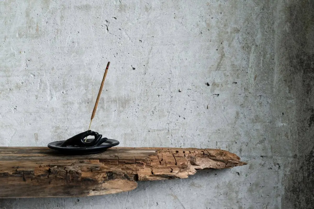 A minimalist incense holder shaped like a hand, crafted from dark ceramic keeping a stick of burning incense, on a grey wall and a piece of wood, representing a mindful Wabi-sabi home gift.