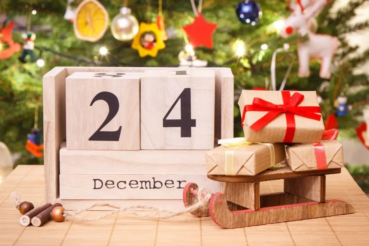 Wooden block calendar showing "24 December" next to wrapped gifts and a Christmas tree.