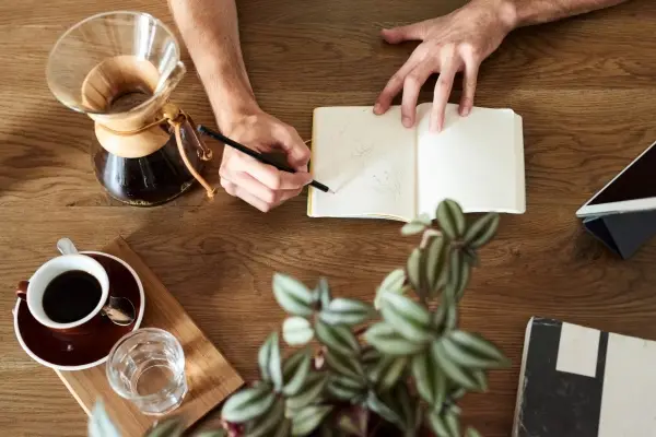 An overhead view of a person writing in a journal on a wooden desk, a simple and creative hygge activity accompanied by coffee and a plant.