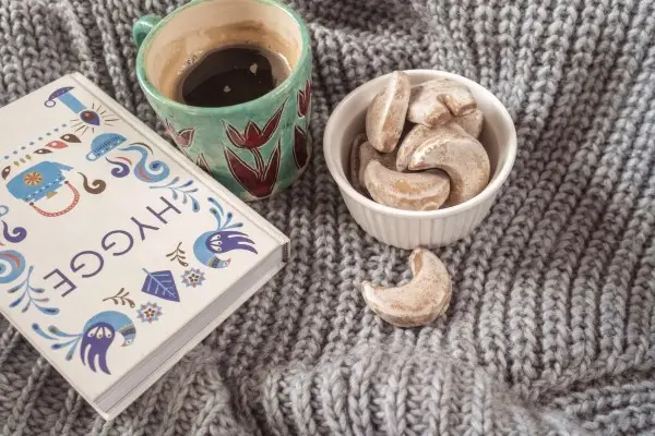 A close-up of a warm cup of coffee and a bowl of crescent cookies resting on a cozy knit sweater, a classic Scandinavian hygge moment.
