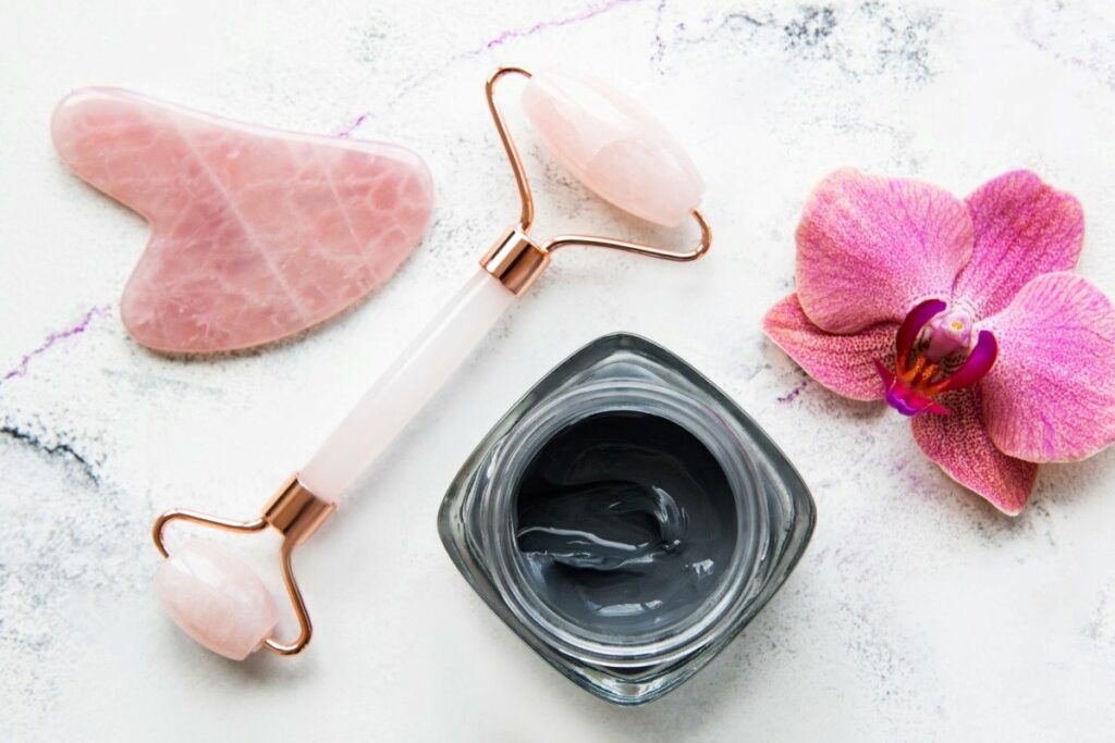 Face jade roller, rose quartz gua sha, and a dark face mask on a marble surface with pink flower.