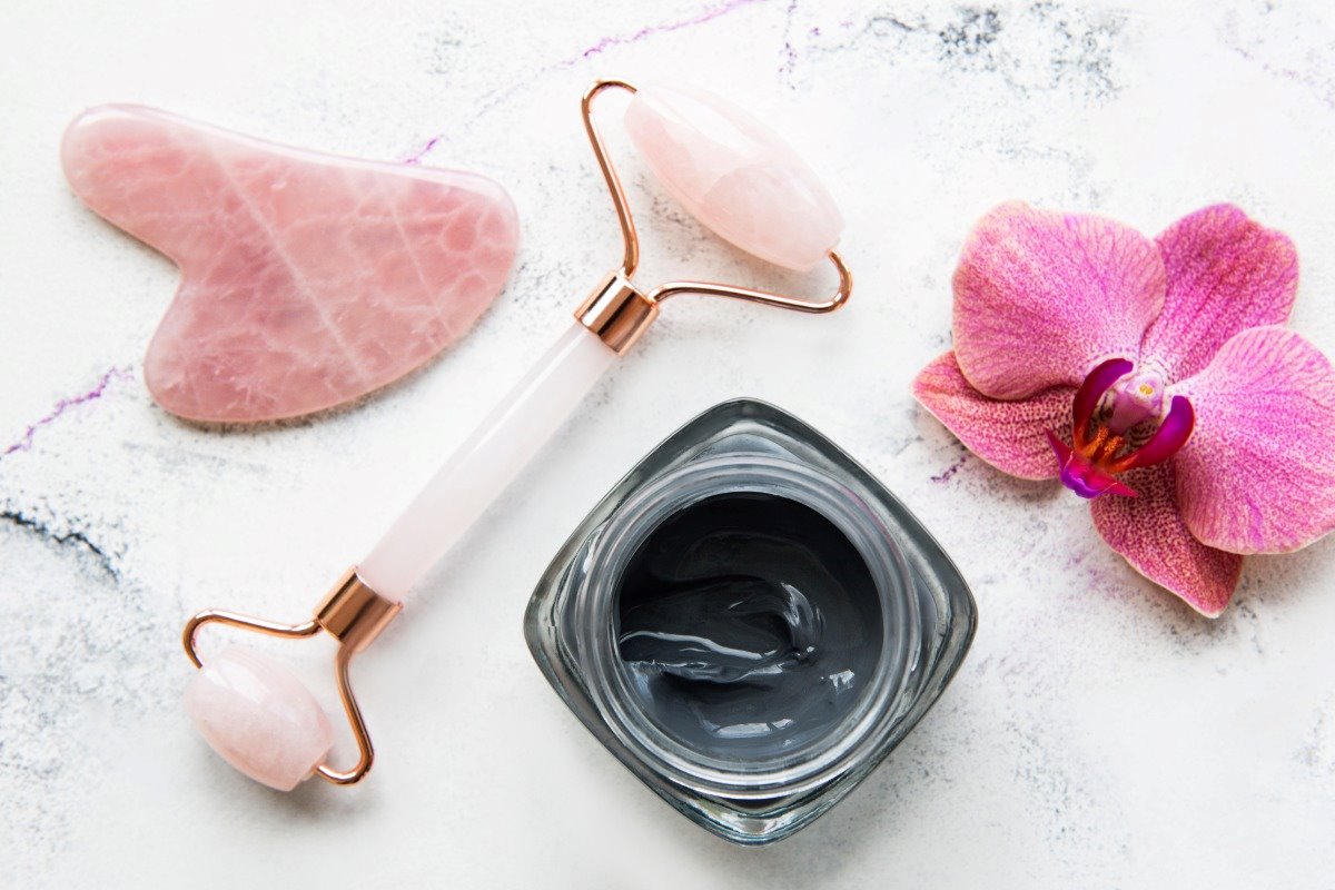 Face jade roller, rose quartz gua sha, and a dark face mask on a marble surface with pink flower.