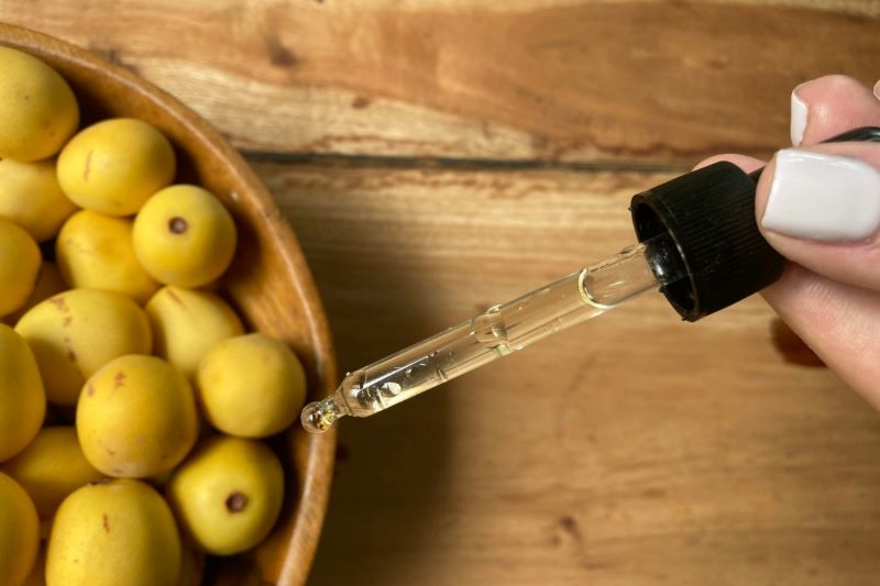 Glass dropper of facial marula oil next to a wooden bowl filled with fresh yellow African marula fruits.