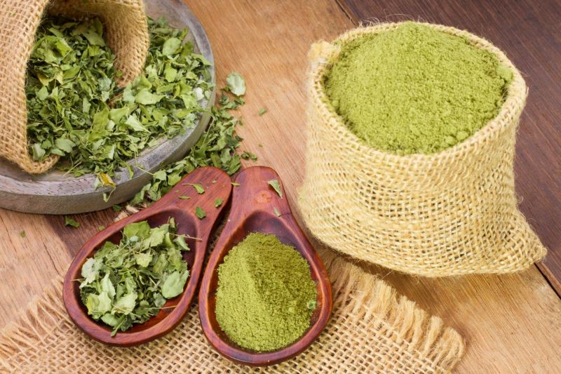Dried African green moringa oleifera powder and leaves arranged in wooden bowls and a burlap sack.