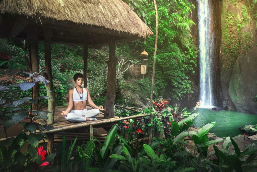 Woman meditating peacefully in a wooden pavilion surrounded by tropical jungle greenery and waterfall in Bali, Indonesia