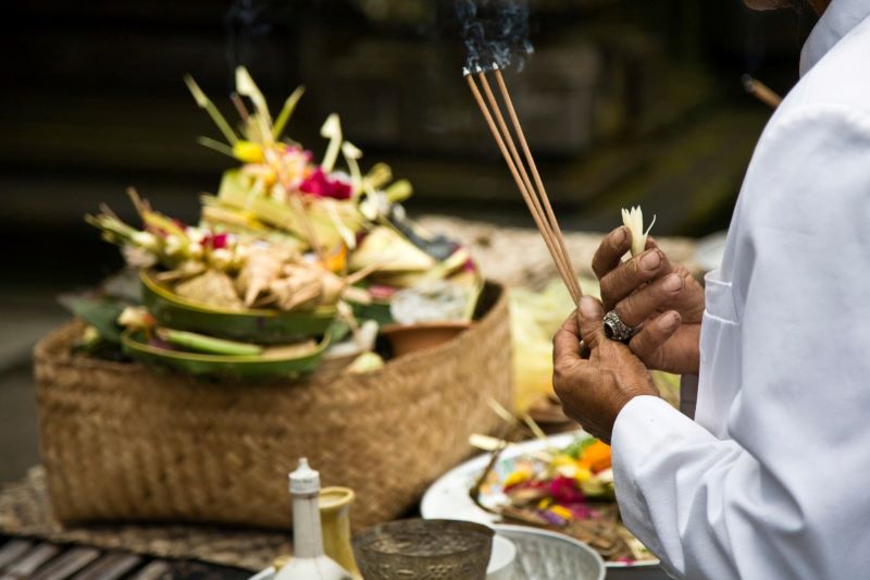 Hands of a man holding burning incense over a traditional Balinese floral offering.